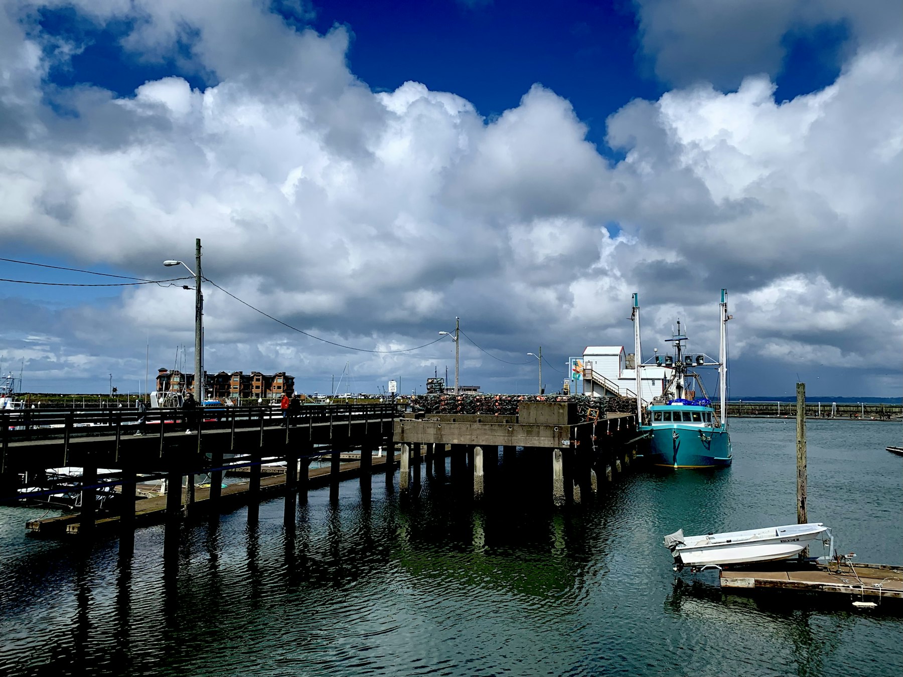 A public wharf on a bright, windy day: a long timber pier with stacked crab traps, a turquoise fishing boat tied alongside, small skiffs on floats, and buildings along the shore behind.